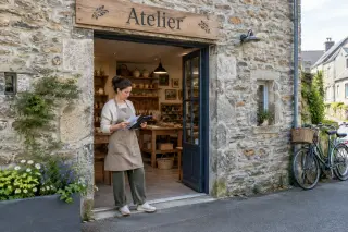 Artisan standing in front of a tidy stone workshop near Quintin, with open door showing shelves of handmade items and bright daylight creating a welcoming atmosphere.